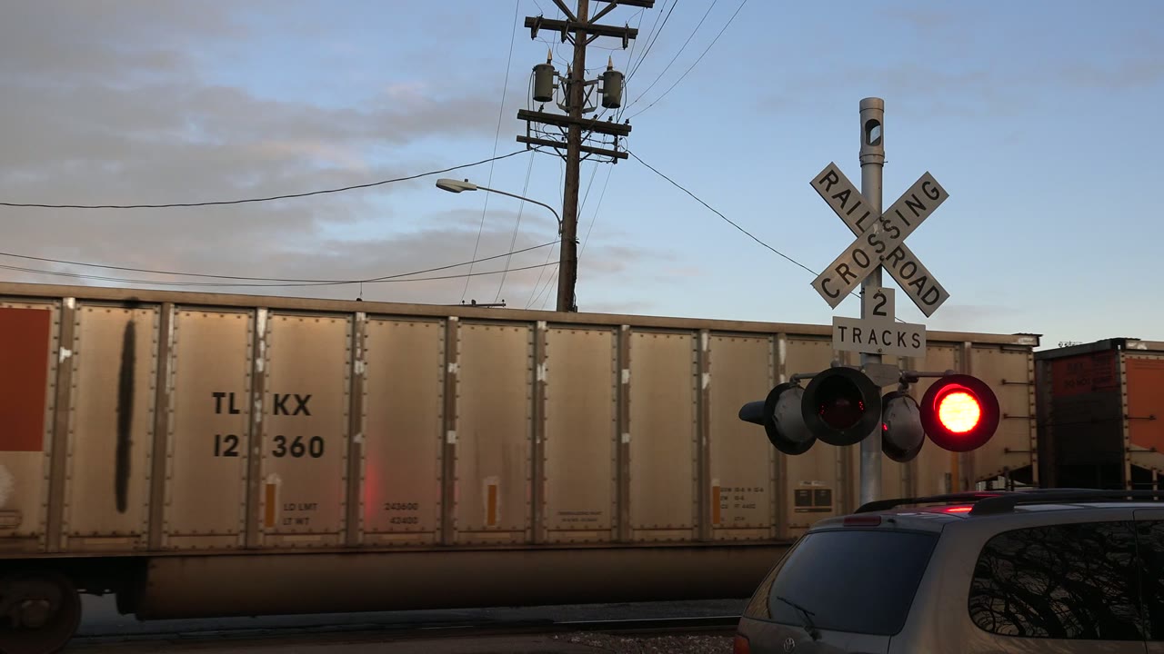 At 0:01 in Westminster, Colorado, the railcar continues across the frame behind the crossbuck and glowing red signal, with utility wires overhead and no visible people.