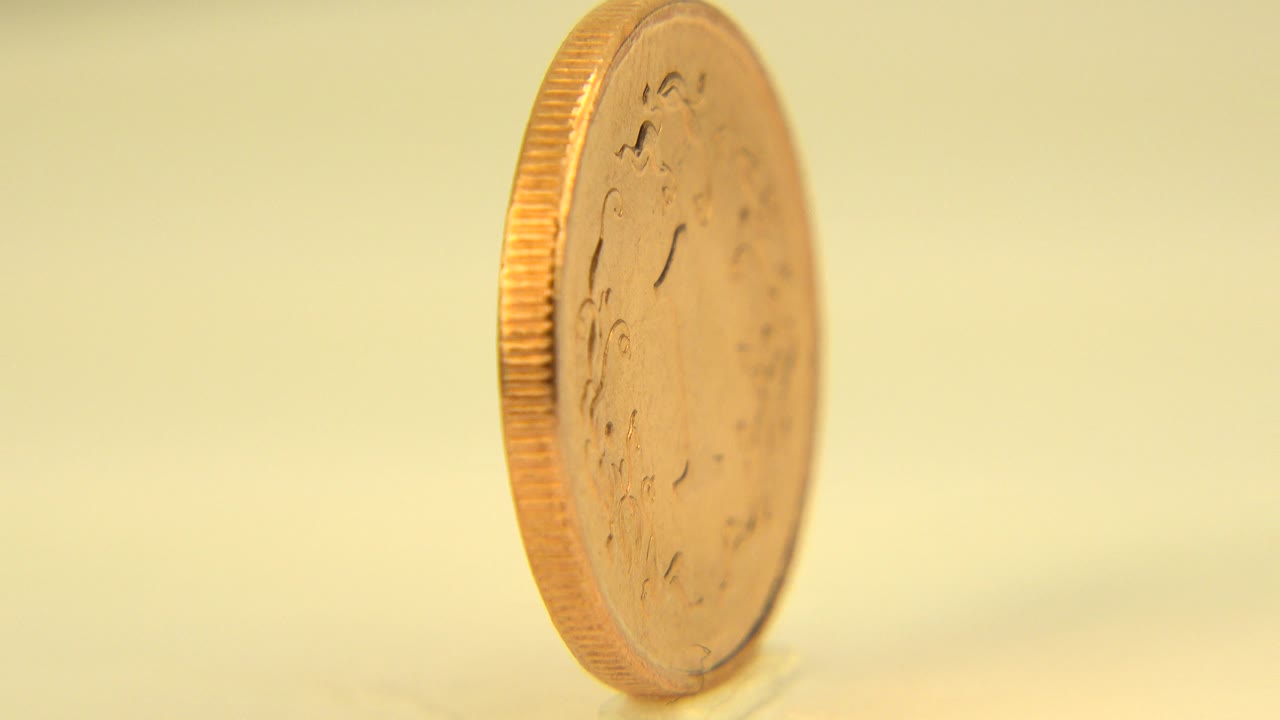 At 0:00 in Westminster, Colorado, a gold-colored coin stands nearly edge-on on a pale reflective surface, with no visible people.