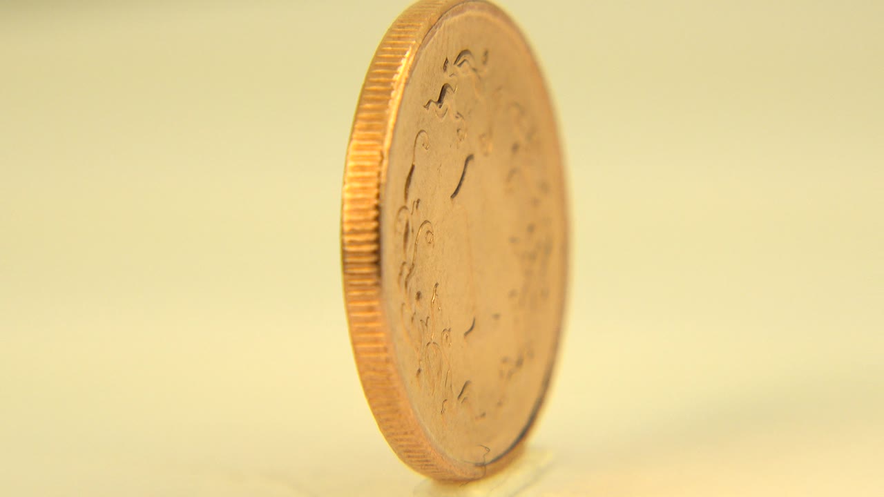 At 0:01 in Westminster, Colorado, the upright gold-colored coin remains mostly edge-on, with a faint embossed face visible and no visible people.