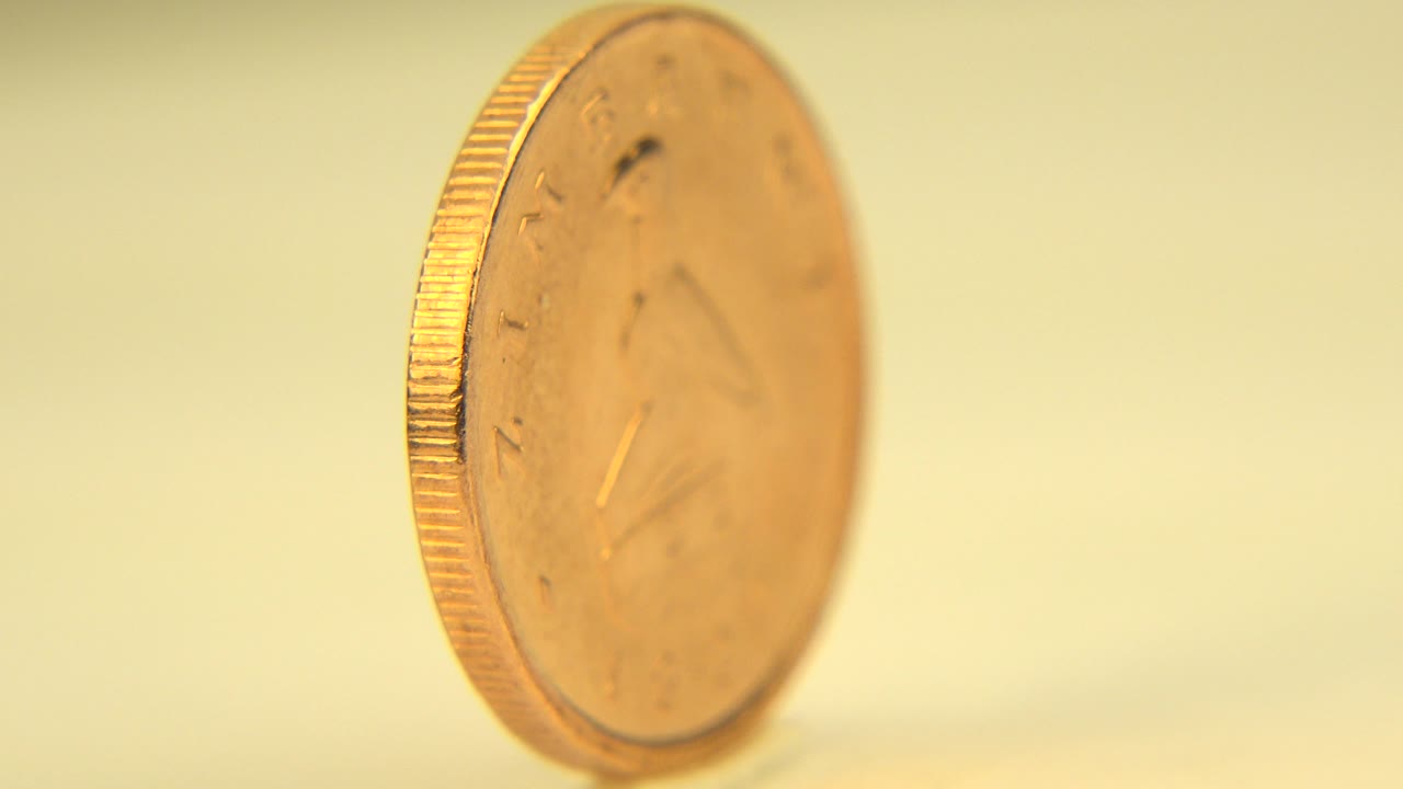 A gold-colored coin spins nearly edge-on on a pale yellow surface in the Westminster, Colorado 2015 clip, with no visible people.