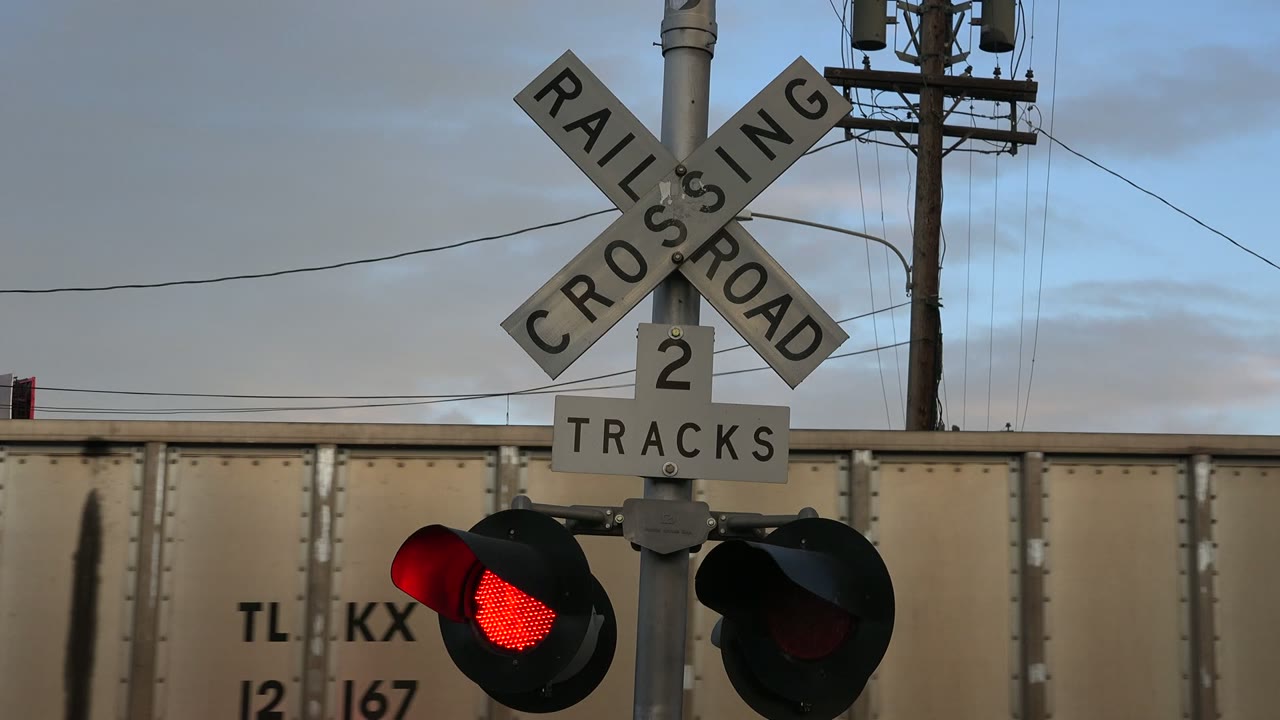 At 0:01 in Westminster, Colorado, the red crossing light glows beneath the crossbuck sign while a beige railcar spans the background, with no visible people.