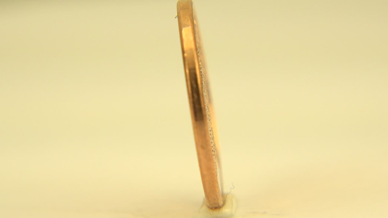 At 0:01, the coin remains nearly edge-on with a small base contact visible on the pale surface and no visible people.