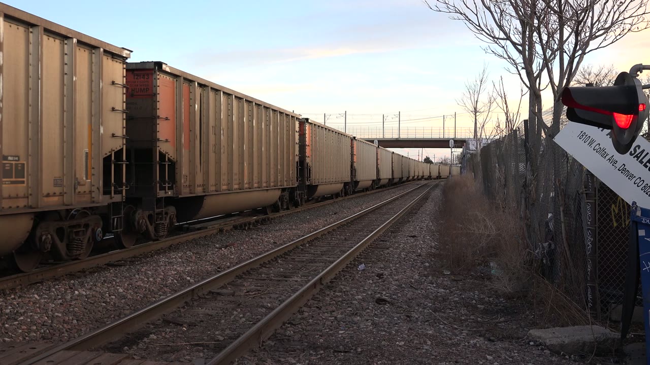 Freight cars continue along the left track in Westminster, Colorado, with rails converging toward an overpass, a red signal light at right, bare branches, and no visible people.