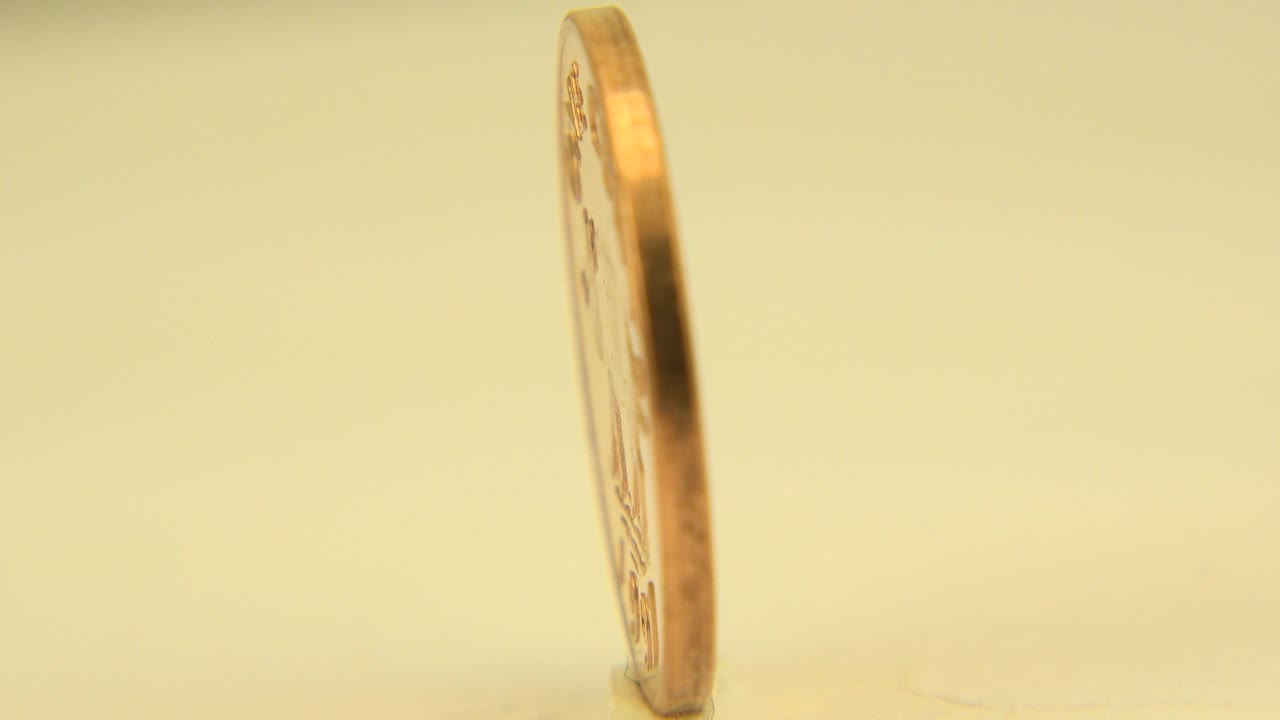 The one-pound coin remains upright in a narrow side view on the pale tabletop, with the Westminster, Colorado clip showing no visible people.