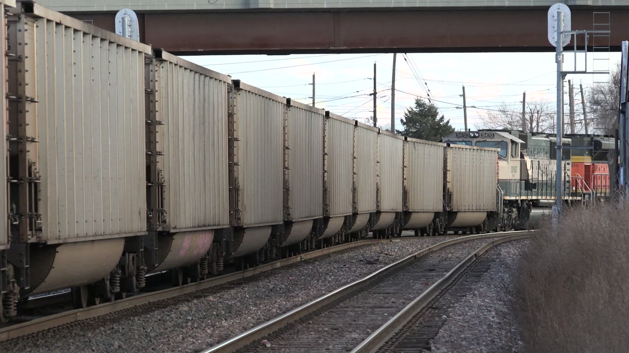 Freight cars run along curving railroad tracks in Westminster, Colorado, with an overpass above, utility poles beyond the rails, brush at right, and no visible people.