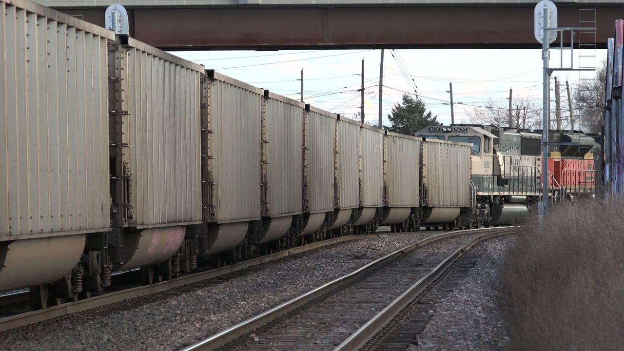 A string of pale freight cars continues through the Westminster, Colorado rail scene, with a red locomotive partly visible behind them and no visible people.