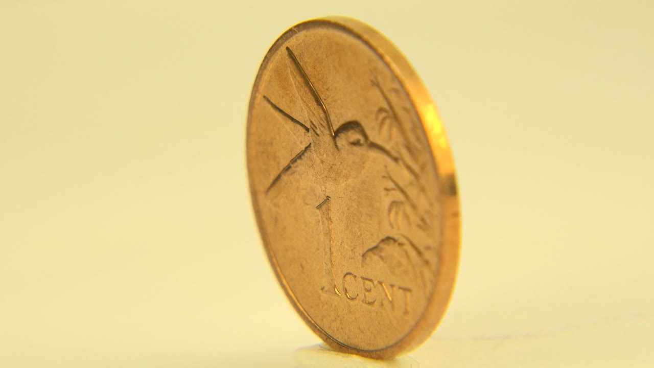 The upright gold coin remains near the center of the frame, its front face and raised markings more visible against the soft yellow tabletop, with no visible people.