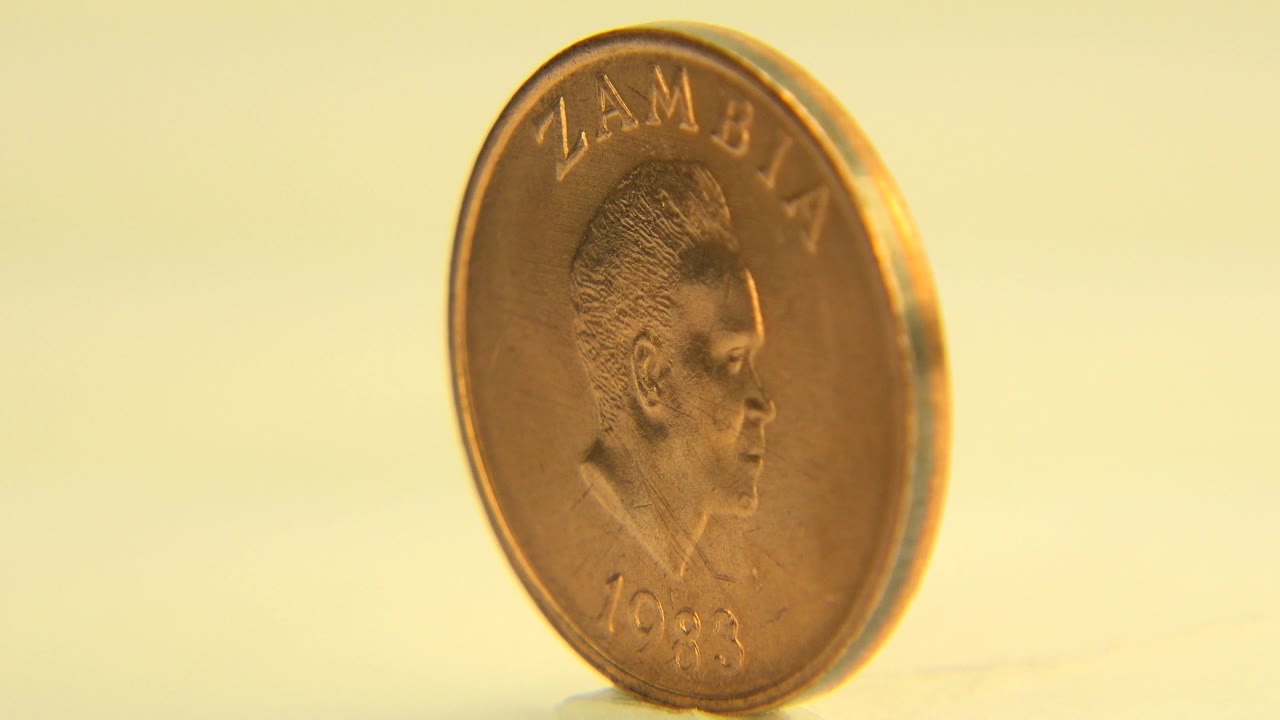 A copper-colored Zambian coin stands upright on a pale reflective surface in Westminster, Colorado 2015 footage, with the portrait side partly facing camera and no visible people.