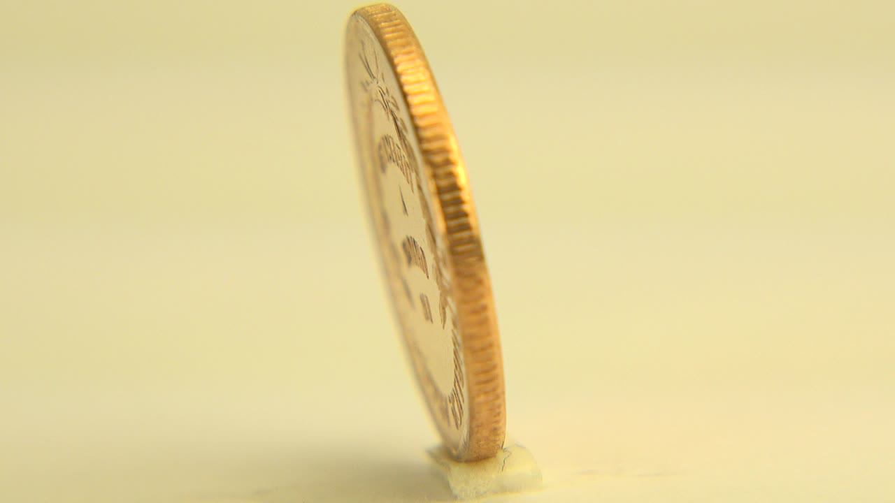 At the start of the Westminster, Colorado clip, a copper-colored centavo coin stands nearly edge-on on a pale yellow surface with no visible people.