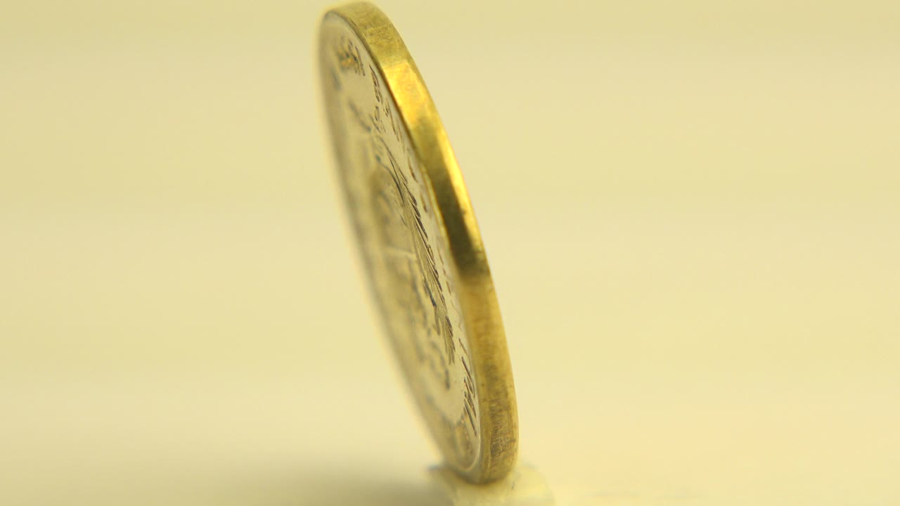 At 0:00 in Westminster, Colorado, a gold-toned one centavo coin is seen mostly from its thin edge on a pale flat surface, with warm light and no visible people.