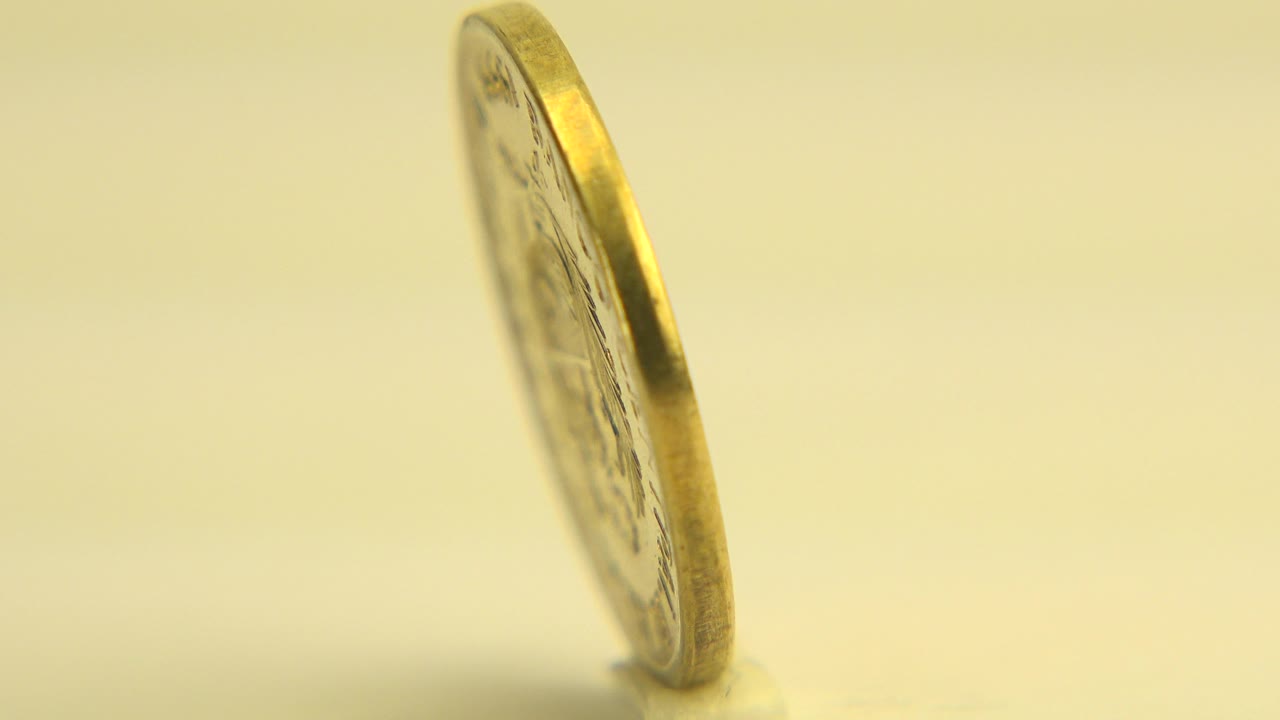 At 0:01 in Westminster, Colorado, the coin remains mostly edge-on with a slight face angle visible, set against a softly blurred yellow background and no visible people.