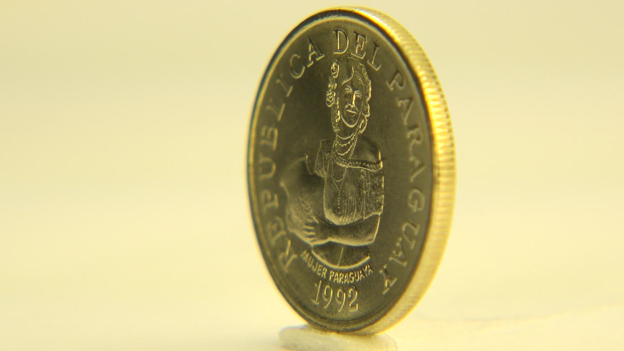 Gold-colored coin standing upright near the left side of the frame in Westminster, Colorado footage, showing a Paraguayan face side under white light with no visible people.