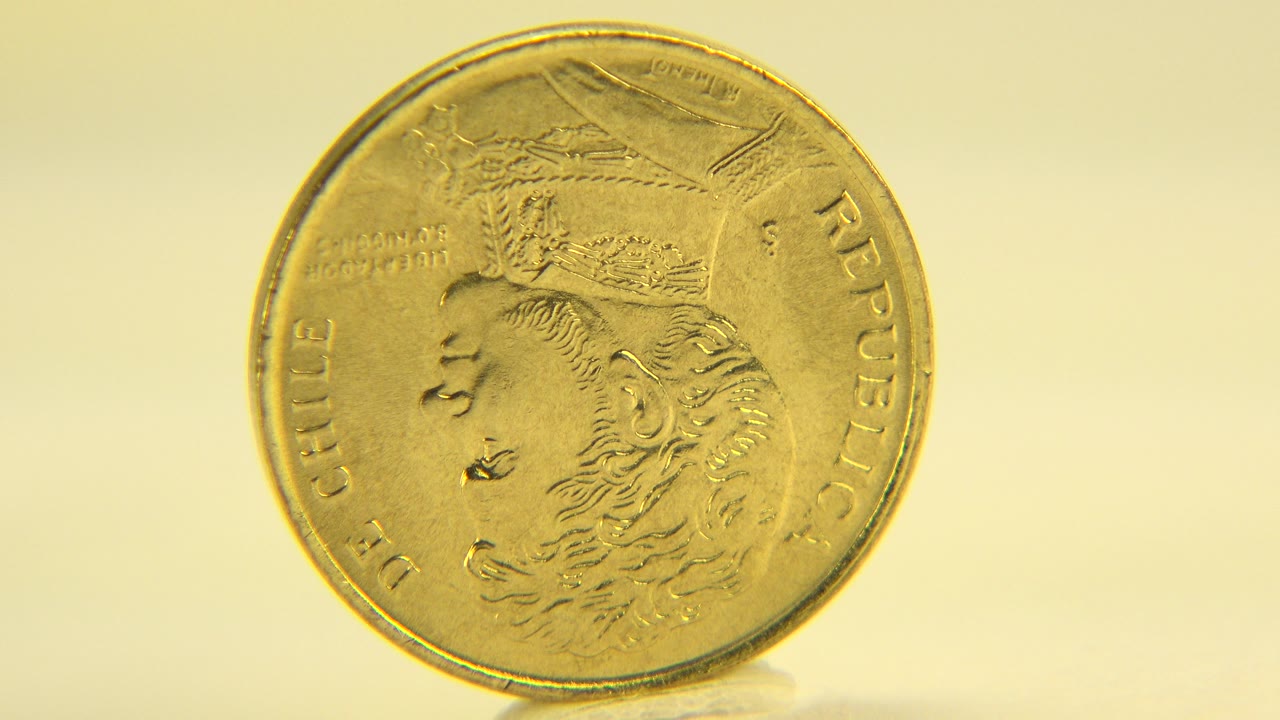 A gold Chilean coin stands nearly face-on on a pale reflective surface in Westminster, Colorado, with warm yellow light and no visible people.