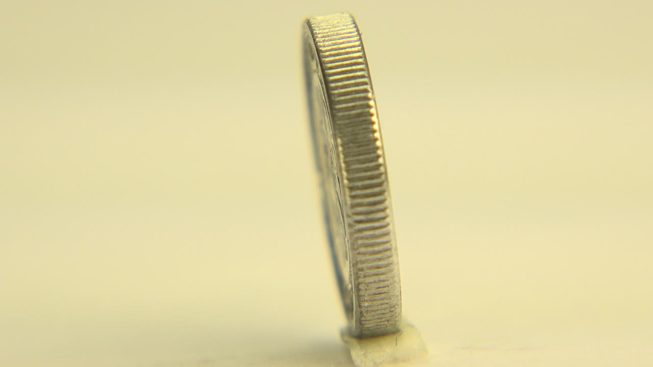 At 0:00, an upright silver-colored coin is seen mostly from its narrow edge on a pale tabletop in Westminster, Colorado, with warm light and no visible people.