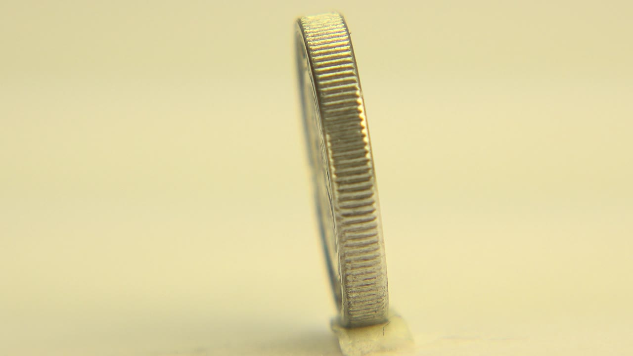 At 0:01, the coin remains almost edge-on in a close low view, with its ridged side catching warm light above the pale tabletop and no visible people.