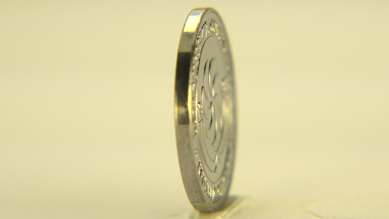 The silver coin remains mostly edge-on in Westminster, Colorado, its rim catching warm highlights against a soft pale background with no visible people.