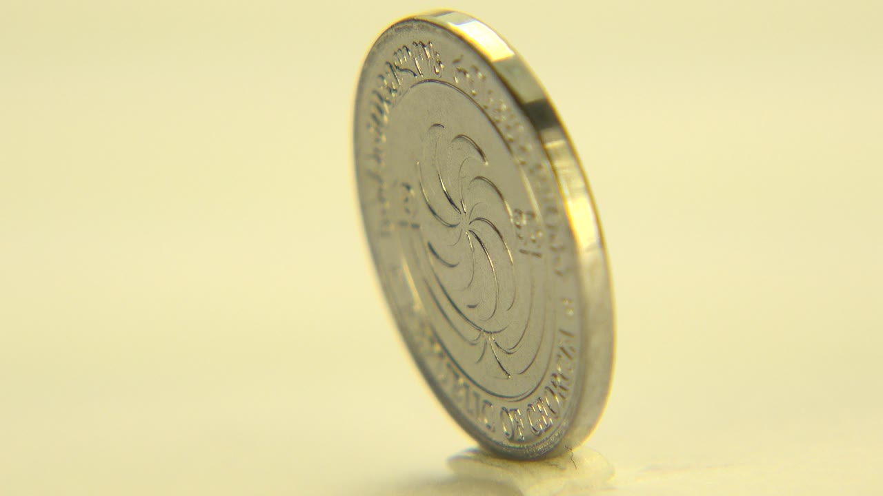 At 0:00, a gold-toned coin stands upright at a left-leaning angle on a pale yellow surface in Westminster, Colorado, with no visible people.