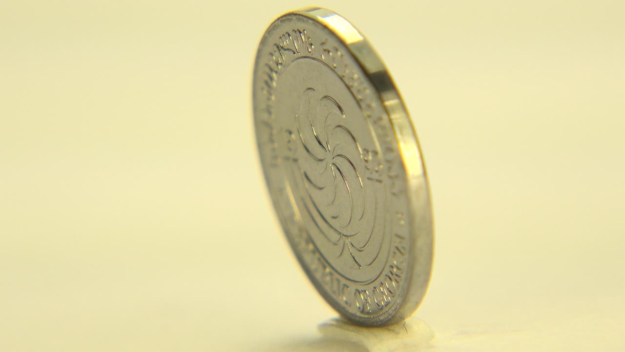 At 0:01, the upright gold-toned coin remains angled on the yellow surface, with a bright rim highlight and no visible people.