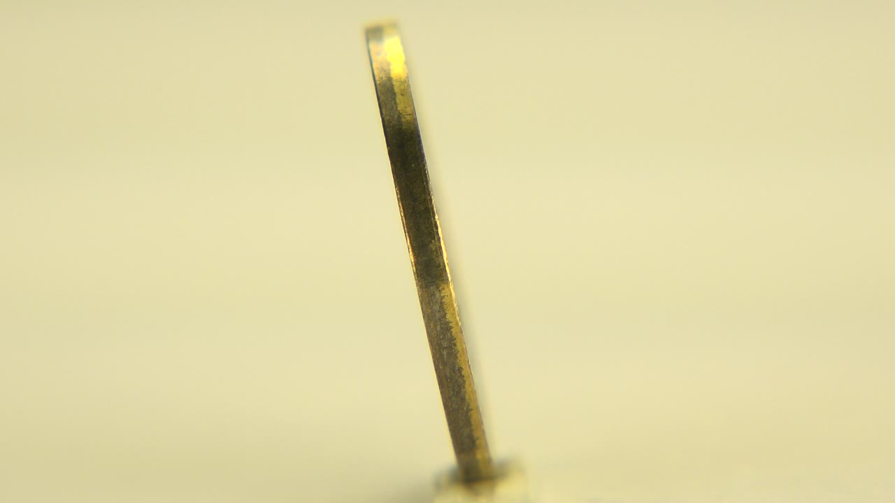 At 0:00 in Westminster, Colorado, a gold-colored coin appears nearly edge-on against a soft yellow background with no visible people.