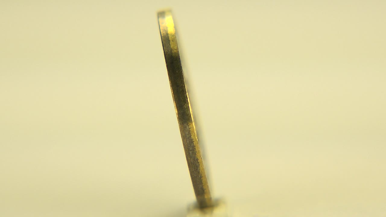 At 0:01 in Westminster, Colorado, the spinning coin remains mostly edge-on, leaning slightly with a blurred yellow background and no visible people.