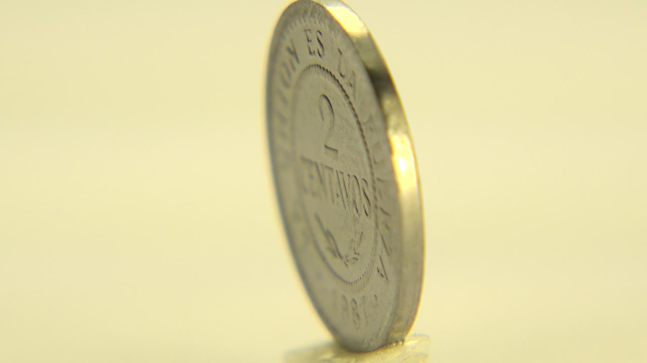 At 0:01, the upright Bolivian coin has turned slightly on the pale surface, catching bright highlights along its rim with no visible people.