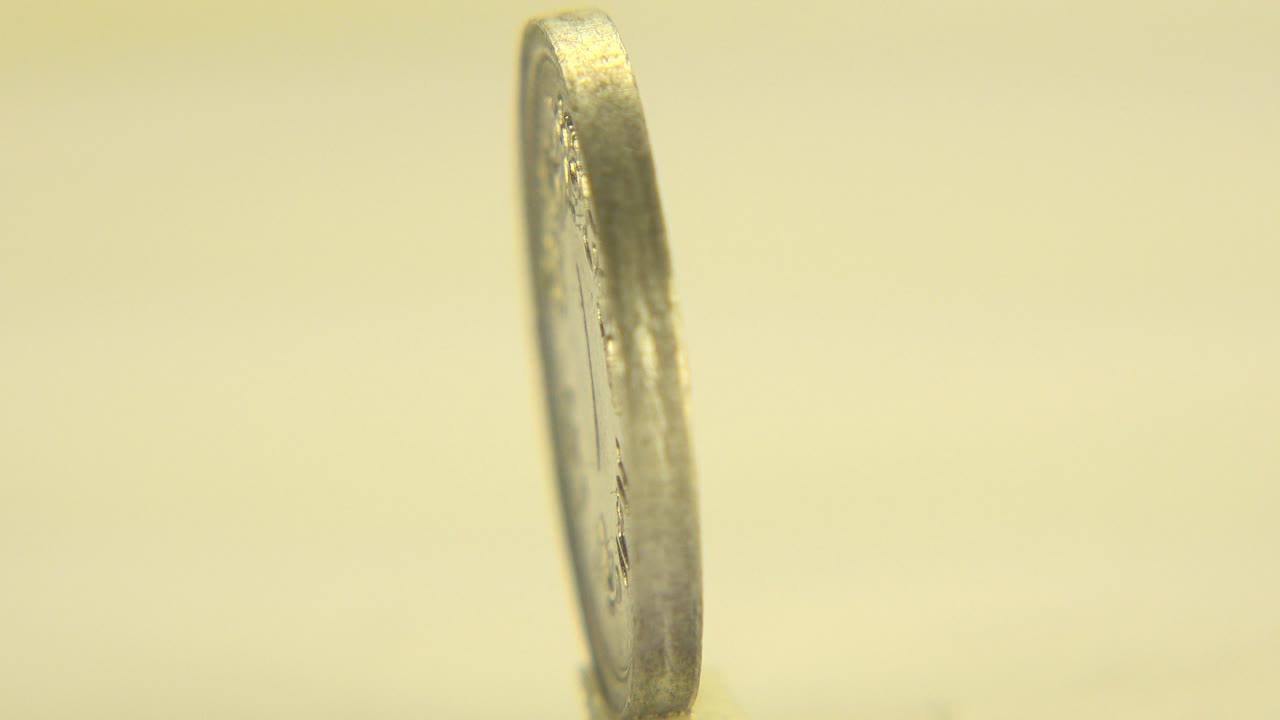 At 0:01, the upper coin remains nearly vertical and the lower coin face is still softly blurred, with warm yellow light across the metallic surface in Westminster, Colorado, and no visible people.