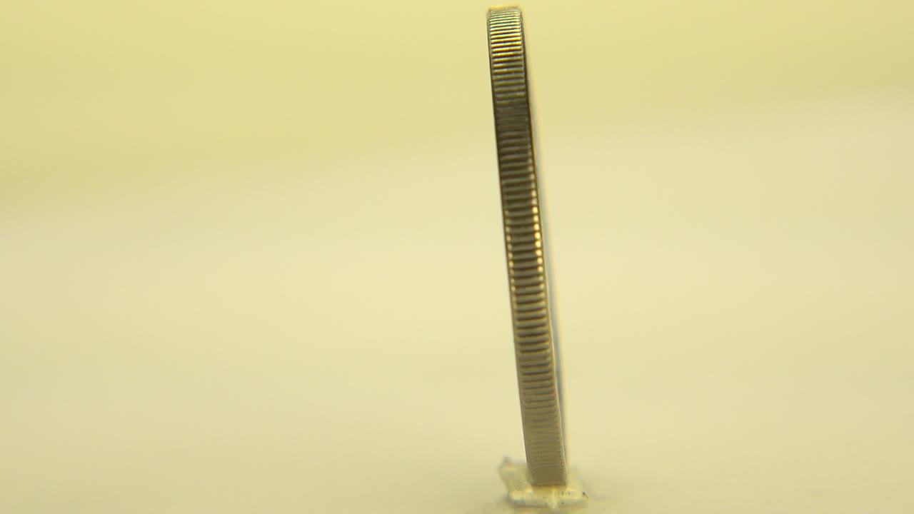 Thin upright coin seen edge-on on a pale yellow surface.