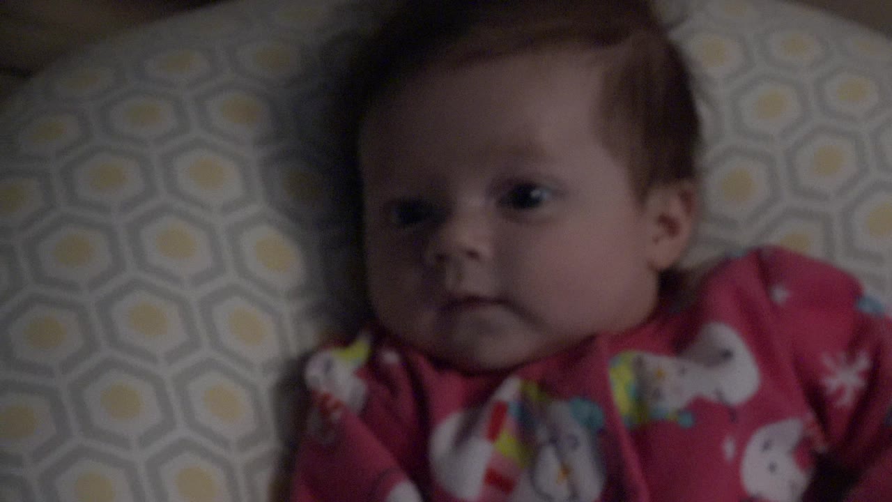 At 0:00, a baby in red printed pajamas lies on patterned bedding in Westminster, Colorado, looking slightly toward the camera in dim indoor light.
