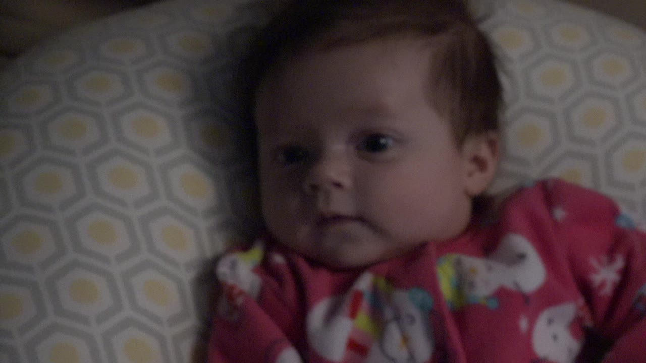 At 0:01, the baby remains on the patterned bed surface, with the face centered more clearly and one red pajama sleeve visible near the lower frame.