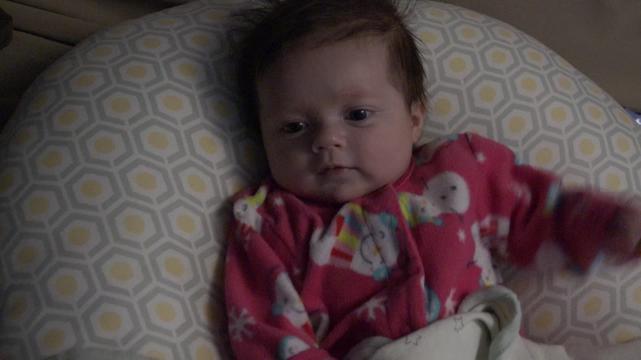 Baby lying on a patterned cushion in dim indoor light.