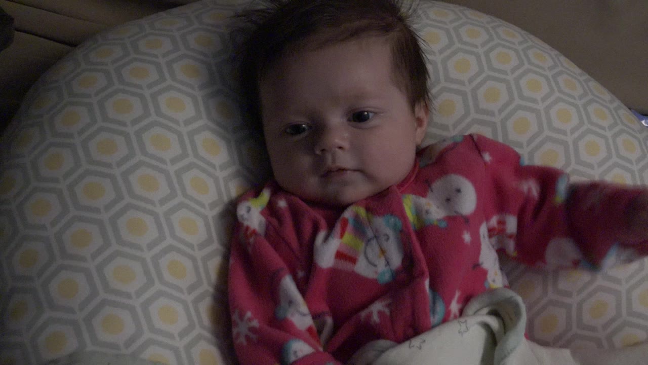 At 0:01, the baby looks toward the camera while one sleeve blurs slightly from movement beside the patterned cushion in Westminster, Colorado.