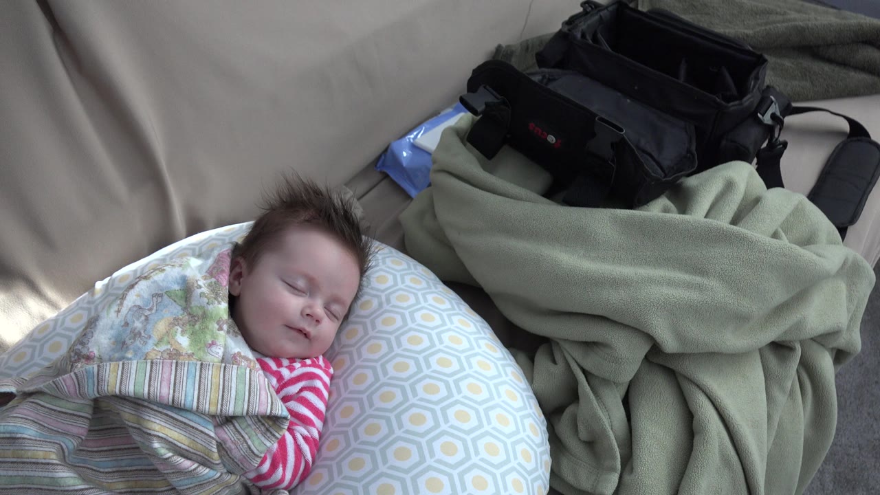 Sleeping baby on patterned pillow beside blankets and a black bag.