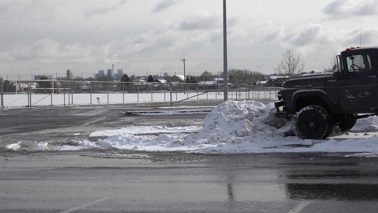 The dark lifted truck sits more fully in view beside a plowed snowbank on a wet Westminster, Colorado street, with fence lines and distant buildings in the background, no visible people.