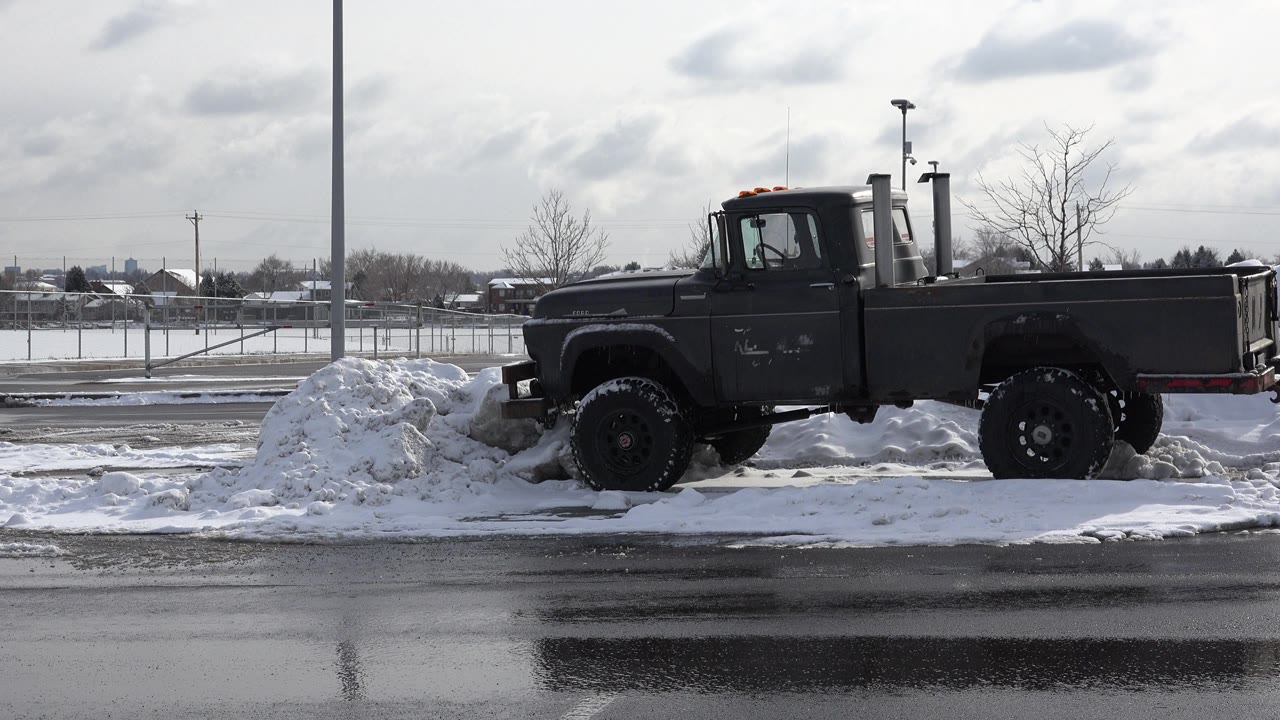 A dark truck is parked side-on beside plowed snow in a Westminster, Colorado lot, with wet pavement in front and no visible people.