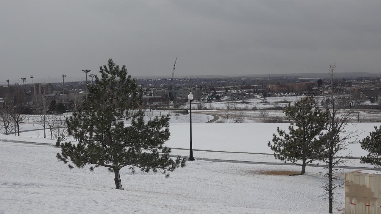 Opening frame of Westminster, Colorado with snow across a sloped park area, evergreen trees in front, a dark path, distant buildings, and no visible people.