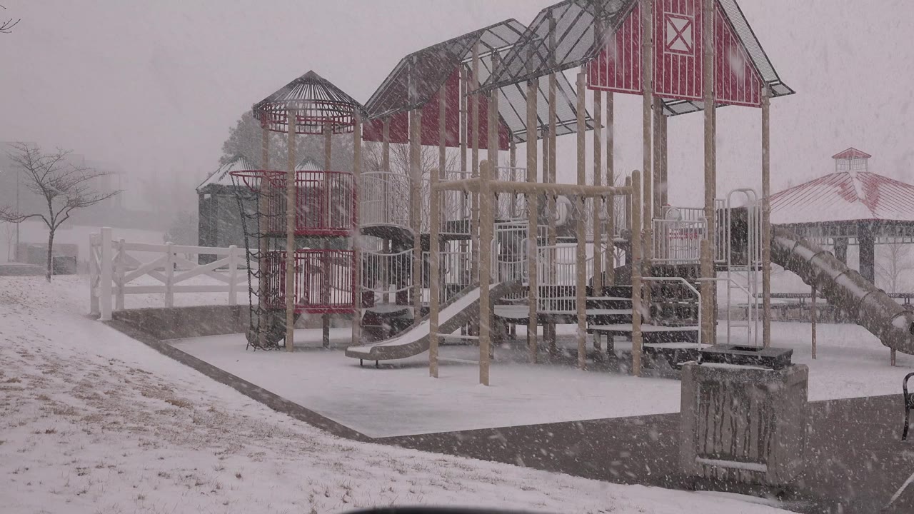 At 0:00, a red and tan wooden playground in Westminster, Colorado stands empty during heavy snowfall, with a trash can in the foreground and no visible people.