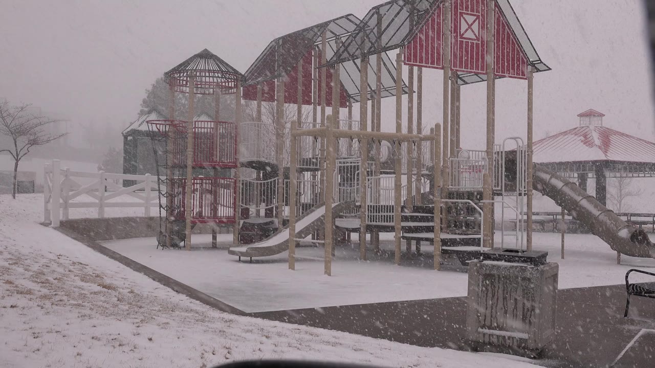 At 0:00, a Westminster, Colorado playground sits empty during falling snow, with red roofed play structures, white fencing, a gazebo, and no visible people.