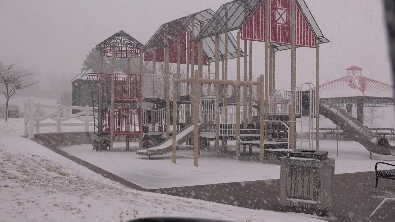 Playground equipment in a snowstorm with gazebo and bare trees in the background.