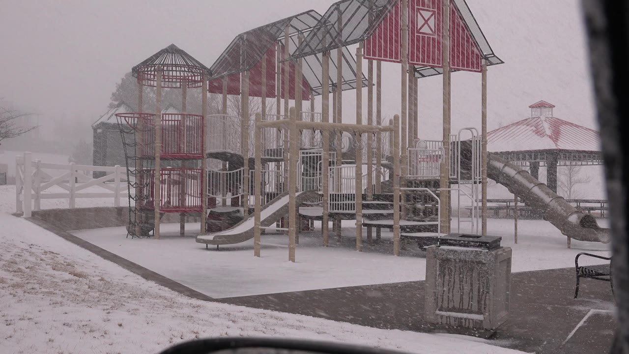 Early frame of an empty Westminster, Colorado playground in falling snow, with red-roofed play towers, slides, a path, fence, gazebo, and no visible people.