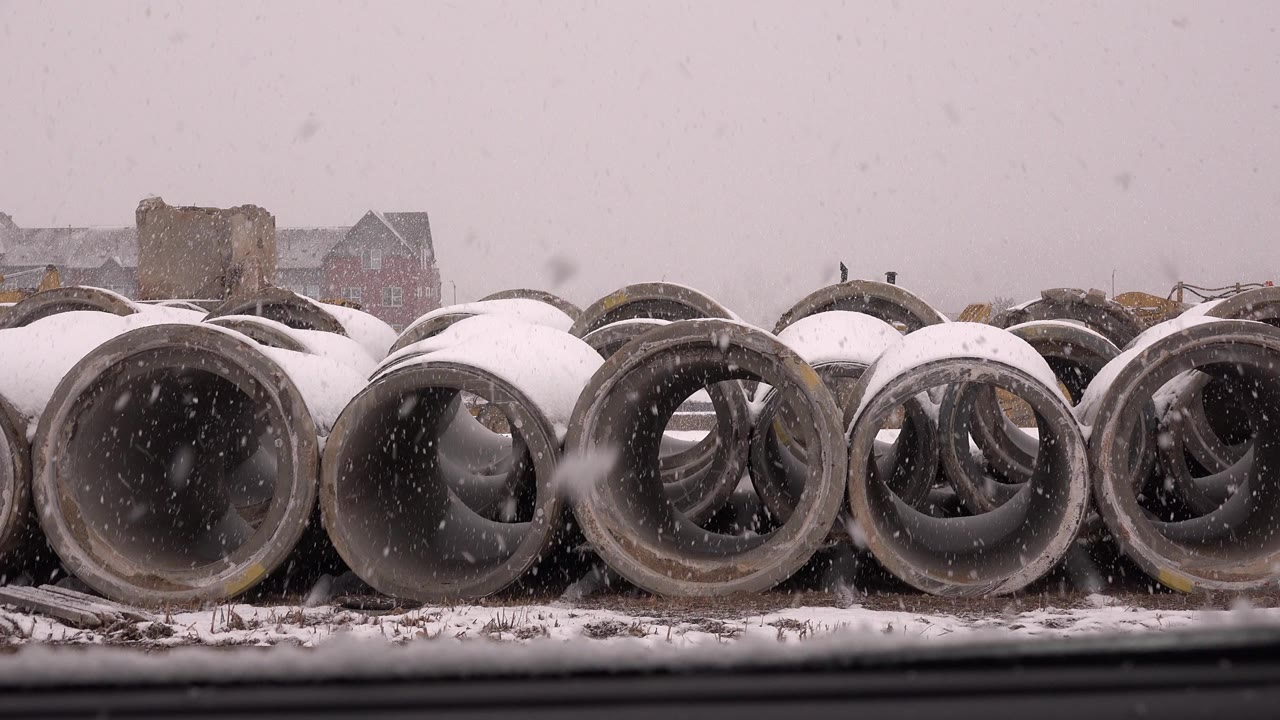 Snow falls over rows of large concrete pipes at a Westminster, Colorado construction site, with distant buildings softened by the weather and no visible people.