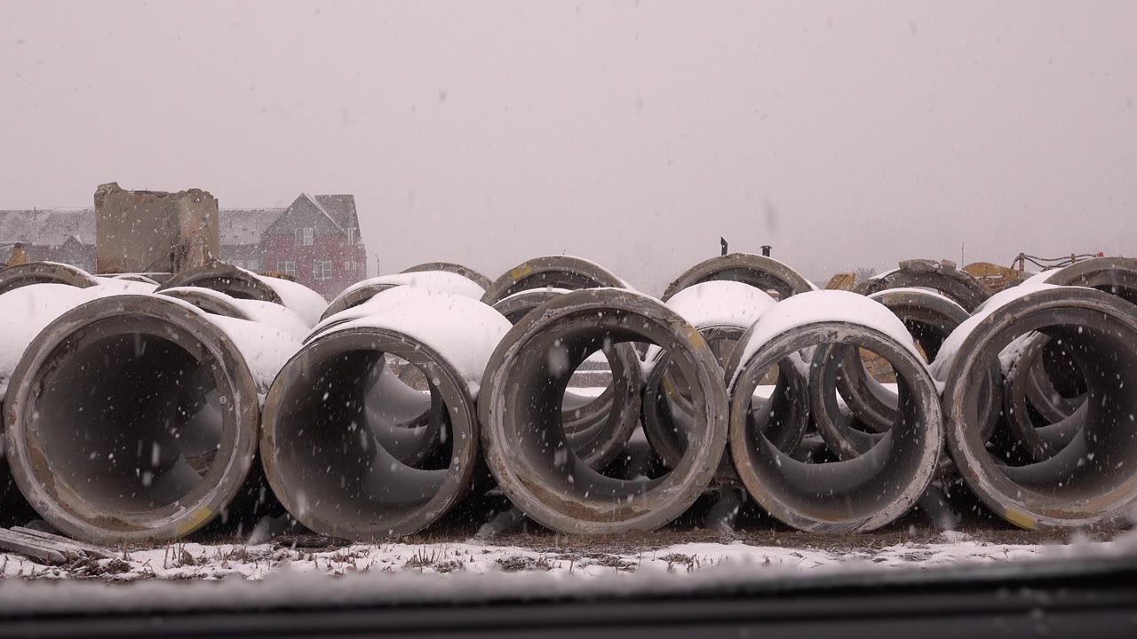At 0:01, circular concrete pipe openings dominate the frame while snow falls across the muted yard and background buildings in Westminster, Colorado, with no visible people.