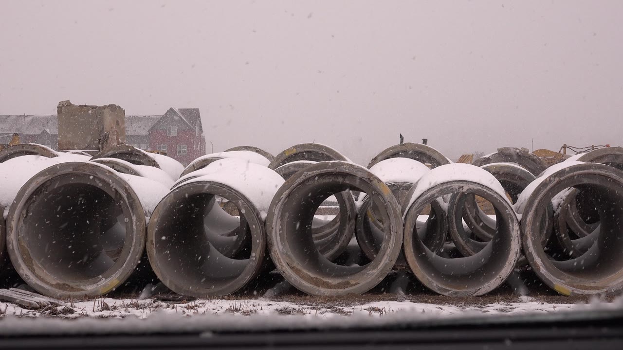 A close ground-level view shows gray concrete pipe rings with white snow caps, muted winter light, distant structures, and no visible people in Westminster, Colorado.