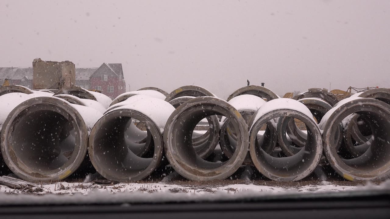 Snow-covered concrete pipes beside a road during snowfall.