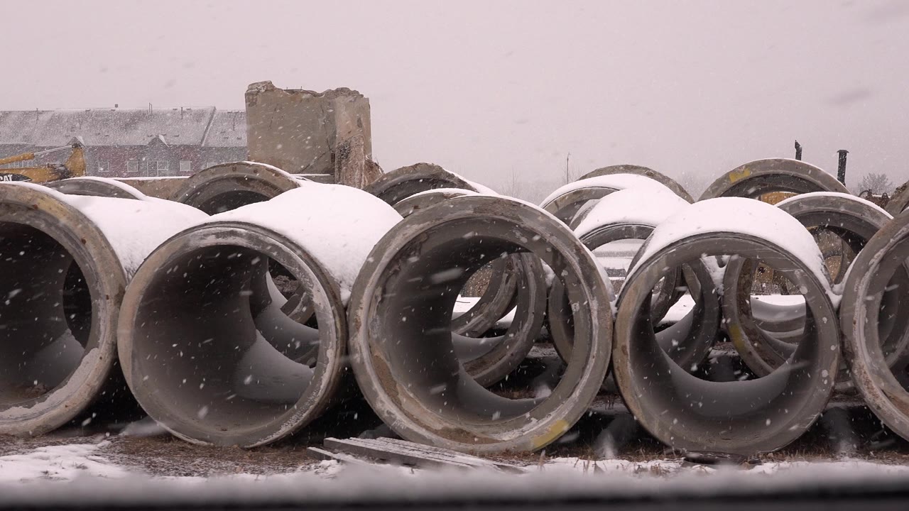 Snow falls over rows of large concrete pipe sections in Westminster, Colorado, with distant buildings and no visible people.