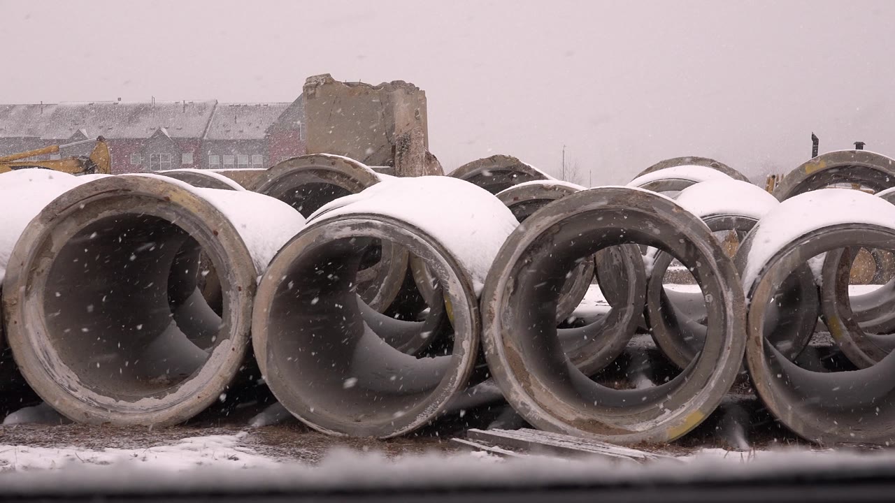 Rows of cement pipe in falling snow with a blocky concrete structure behind.