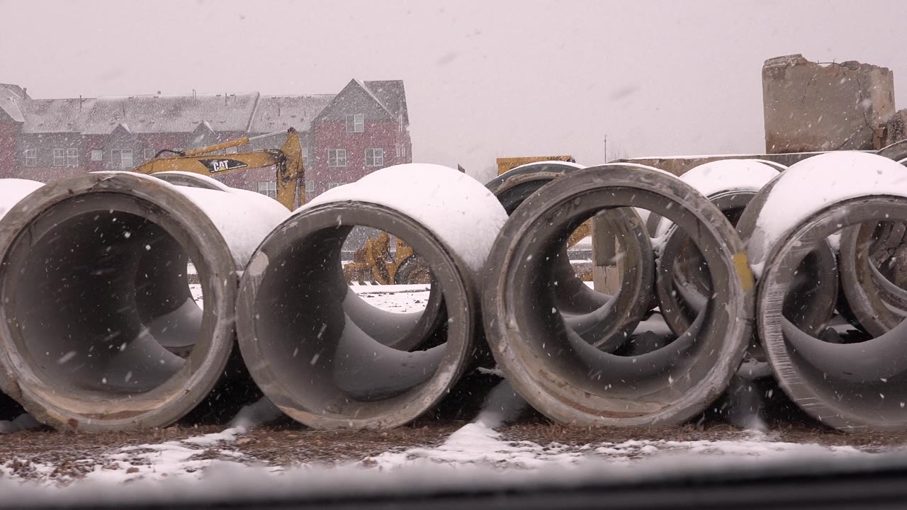 Snow-covered concrete pipes at a construction site with yellow excavators behind them.