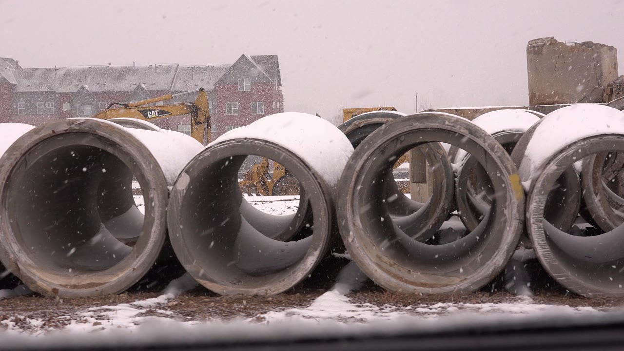 Snow falls over a row of round concrete pipe sections in Westminster, Colorado, with an excavator arm partly visible behind the pipes and no visible people.
