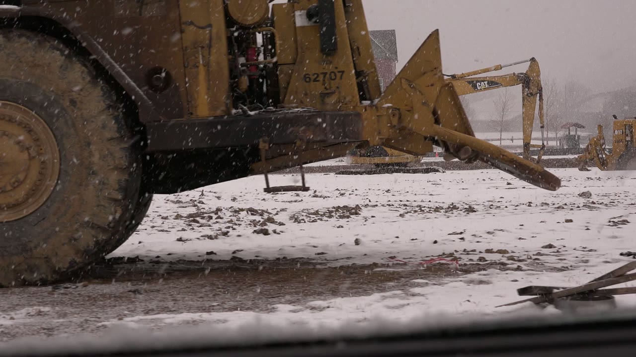 A large muddy tire dominates the foreground as the yellow construction vehicle passes across snowy ground in Westminster, Colorado, with no visible people.