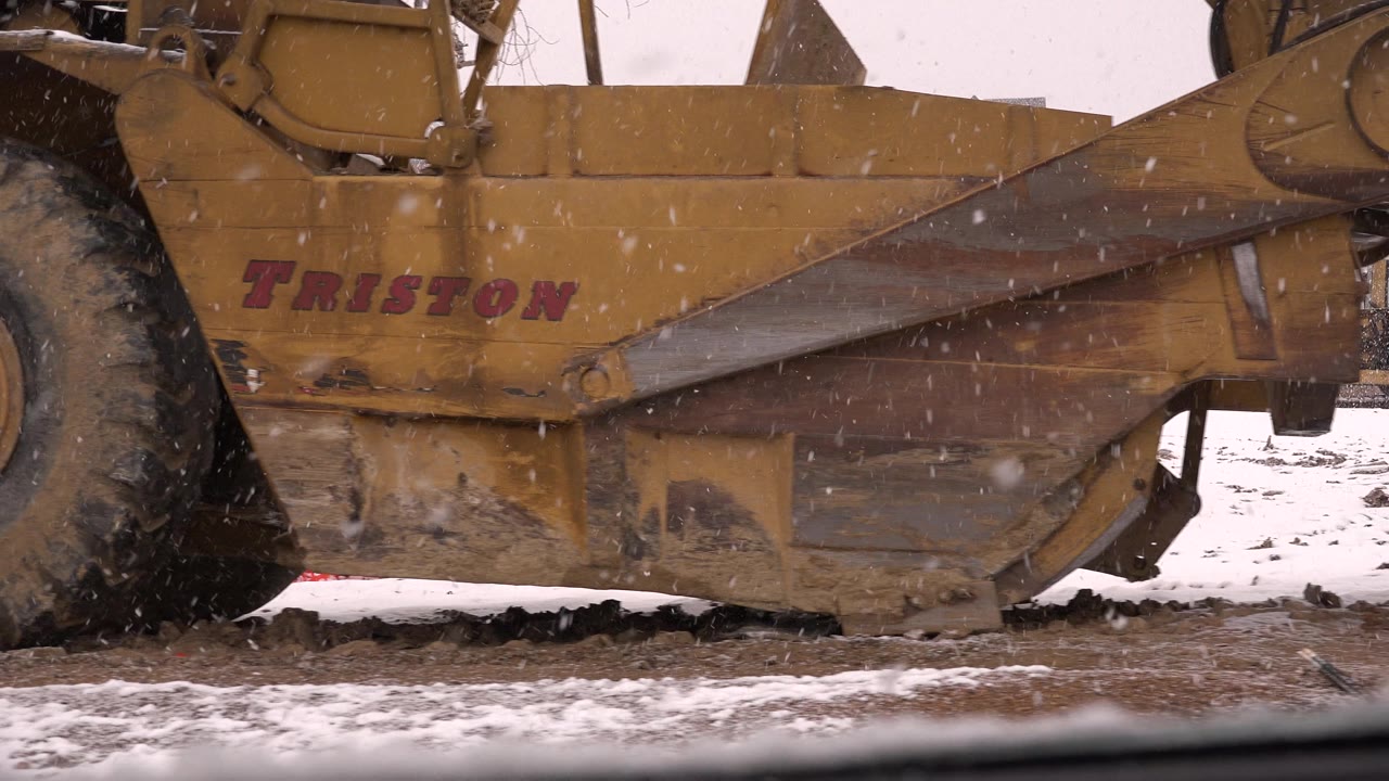 The yellow Triston-marked scraper tractor fills the foreground in Westminster, Colorado, with snow falling over muddy ground and no visible people.