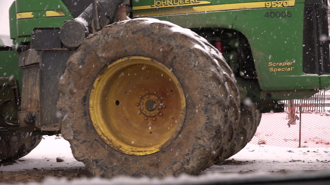 Large muddy tractor wheel beside a green machine body in Westminster, Colorado, with yellow trim, falling snow, and no visible people.